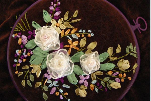 Mit Liebe zum Detail ist diese Stickarbeit mit zarten Stoffblumen zusätzlich verziert. im Hintergrund Handstickerei auf dunklem Grund.